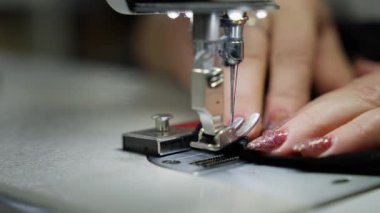Siyah iplikle dikiş yapan bir dikiş makinesi ayağının yakın çekimi. Kadınların parmakları dikiş yapmak için kumaşı hareket ettirir ve iğneleri malzemeden çıkarır. Giysi imalatı, terzilik atölyesi.