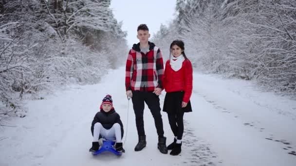 一个年轻的夫妻家庭在冬天的公园里散步 森林里的雪橇上带着一个孩子 年轻的父母和一个孩子在冬季公园里散步 — 图库视频影像