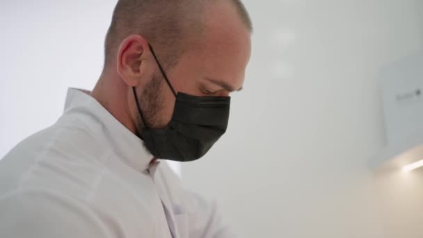 白いコートを着て黒い顔のマスクを着用した男性医師は 集中して見下ろし 臨床的な設定を示唆しています — ストック動画