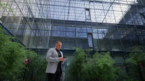 ビジネスマンは意図的にスマートフォンを見て 緑豊かな植物の背景に対してデジタル時代の彼のビジネスを管理します — ストック動画