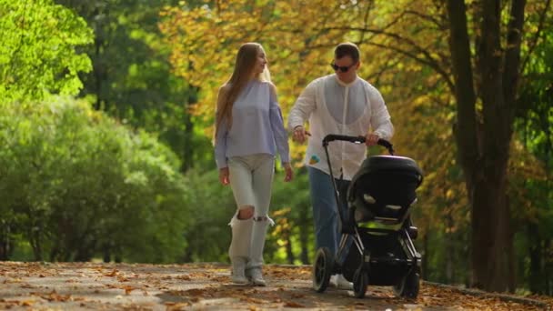 愛するカップルは 公園の路地に沿ってベビーキャリッジを歩いています 屋外で新生児と歩く白人男性と女性 平和を楽しむ — ストック動画