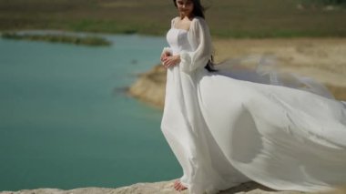 Göl sakinliğini kucaklayan, dalgalı beyaz elbiseli bir gelin dingin anın tadını çıkarmak için duruyor, peçesi ve elbisesi engebeli arazinin yumuşak bir kontrastı.