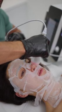 Kapsamlı bir yüz tedavisinin yakın çekimi, kozmetik uzmanının maske takmış bir hastanın yüzünde bir cihaz kullanması, cilt sağlığına çift yönlü yaklaşımı vurguluyor..