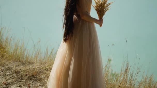一个女人 泰然自若 泰然自若地站在平静的蓝水边 凝视着远方的地平线 手里拿着一丛簇小麦 象征着自然的恩惠 — 图库视频影像