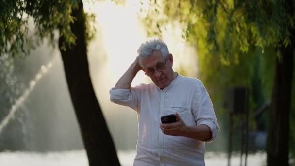Ηλικιωμένος Άνθρωπος Στοχαστική Στάση Smartphone Στο Πάρκο Στο Ηλιοβασίλεμα — Αρχείο Βίντεο