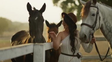 Nazik beyaz elbiseli bir kadın onlarla iletişim kurarken atları okşar..