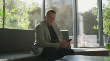 Düşünceli bir profesyonel, akıllı telefonuyla meşgul. Güneşle ıslanmış bir salonda oturuyor. Zeminden tavana kadar yeşil manzaralı..