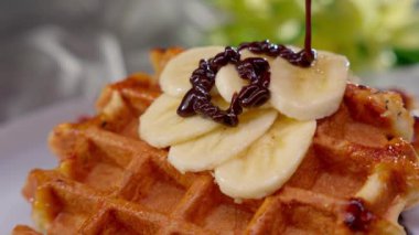 Klasik Belçika waffle 'ı muz mermileriyle kaplı ve siyah çikolata şurubuyla süslenmiş, el değmemiş beyaz tabakta servis ediliyor..
