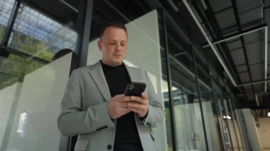 Elinde akıllı bir telefonla, modern bir ofis avlusunun geometrik zarafeti tarafından çerçevelenmiş dijital dünyada bir yönetici ayakta duruyor..