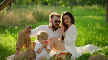 Kadın ve erkek karpuz tadarlar, çocuk da yer, aralarında otururlar..