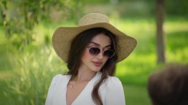 Yaz Şapkalı Zarif Kadın. Bayan hasır şapkasını düzeltiyor, güneşli parkta gülümsüyor..