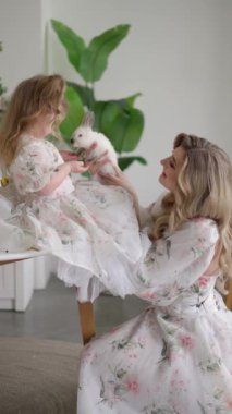 Çiçekli elbiseli anne ve küçük kız küçük beyaz bir tavşan tutarlar, sıcak aile ortamında birbirleriyle nazik bir şekilde etkileşirler..