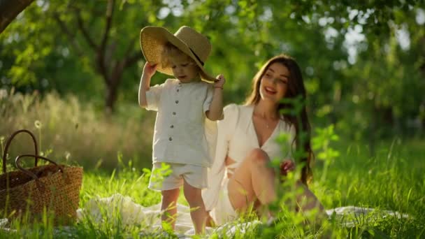 広がった帽子の子供は草を探検し 母親は笑顔で時計をしています — ストック動画