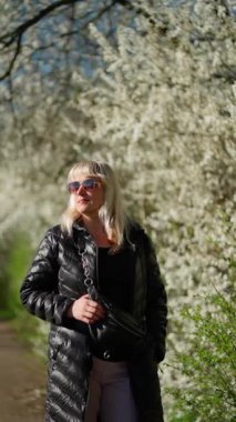 Güneş gözlüklü ve siyah ceketli şık bir kadın çiçek açan ağaçlarla parkta yürüyor..