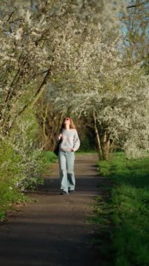 Açık renk kazaklı ve kot pantolonlu genç bir kadın park yolunda yürüyor, beyaz bahar çiçeklerinin arasında güneşli bir günün tadını çıkarıyor..