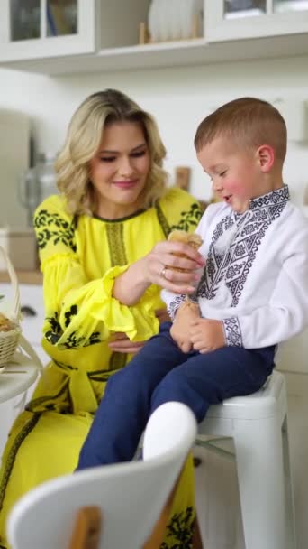 ウクライナの装飾を持つ黄色い服の女性は笑っている 小さな鳥を抱えている 彼女の刺繍されたシャツの彼女の息子は彼女の隣に座っている — ストック動画
