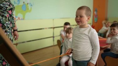 Çocuk aktif olarak sınıf aktivitelerine katılır, akran grubunun önünde durur. Beyaz kazaklı neşeli çocuk elinde sopayla tahtanın önünde duruyor..