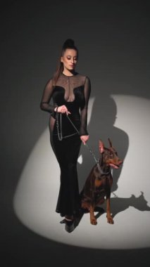 Siyah elbiseli manken karanlıkta Doberman 'a tasma takıyor. Zarif kıyafetli bir kadın, köpeğe tasma takıyor..