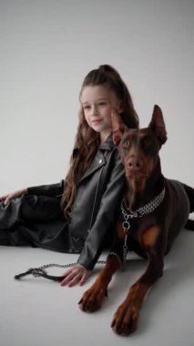 Siyah takım elbiseli çocuk modeli köpeğiyle birlikte beyaz arka planda oturuyor..