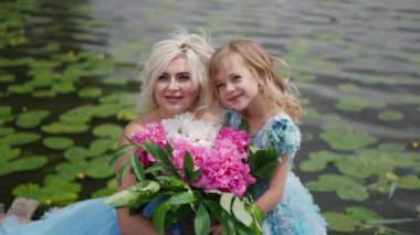 Anne ve kız su zambaklarıyla kaplı göletin yanında büyük bir buket pembe ve beyaz çiçeklerle poz veriyorlar. Güzel elbiseler giymişler, yüzleri gülücüklerle parlıyor, yansıtıyorlar.