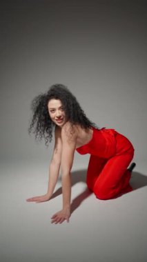 Kıvırcık siyah saçlı, parlak kırmızı elbiseli kadın yere dizlerinin üstüne çöküp kameraya gülümsüyor..