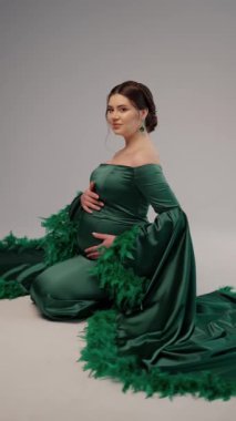 Kucağında tüyleri olan zarif yeşil elbiseli hamile bir kadın. Saçlarını örüyor, yeşil küpeleri var, elleri nazikçe karnına sarılıyor, ifadesi sakin ve memnun..