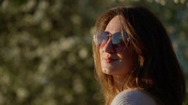 Güneş gözlüklü kadın çiçek açan ağaçların arka planına karşı güneş ışığından hoşlanır. Kendini parlak güneşten korumak için elini kaldırdı. Bahar ve rahatlama atmosferi yarattı..