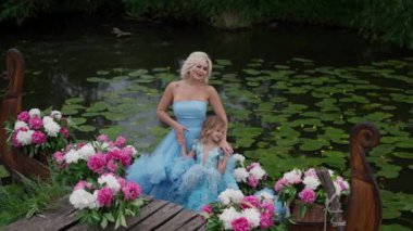 Anne ve kızı resimli gölün arka planında mor elbiseler içinde zambaklarla poz veriyorlar. Pembe ve beyaz çiçeklerin süslemeleri romantik ve şenlikli bir hava katar..