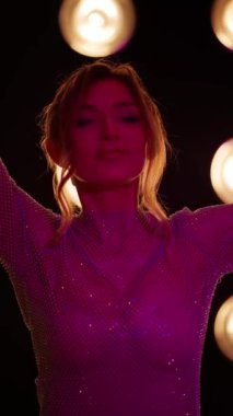 Parlak elbiseli kadın, mavi ışık altında, arka plandaki karanlık stüdyo alanının parlak ışıklarıyla poz veriyor. Şık ve göz alıcı görünüyor..