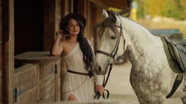 Şık elbiseli kadın ahırın yanında duruyor, yanında da eyerli beyaz at var. O, örgü elbise ve şapka giyiyor, rahat ve memnun görünüyor..