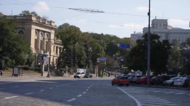 Tarihi binalarla şehir caddesi manzarası. Solda dekoratif elementlerle dolu eski bir kütüphane var ve sağda modern arabaları ve yeşil ağaçları görebilirsiniz, yol kaldırım taşlarıyla döşeli..