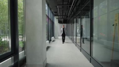 Takım elbiseli bir adam modern ofis binasının koridorunda yürüyor. Yeşil bahçeye bakan şeffaf cam duvarlar tasarımın stilini ve modernliğini vurguluyor.