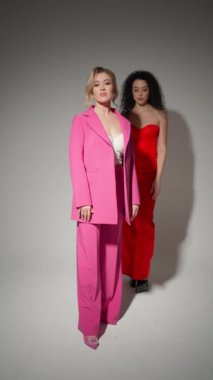 İki kadın stüdyo fotoğraf çekiminde poz veriyor, biri pembe takım elbiseli, diğeri kırmızı elbiseli. Özgüvenleri ve tarzları modaya uygun imaj ve kadınlığı vurguluyor..