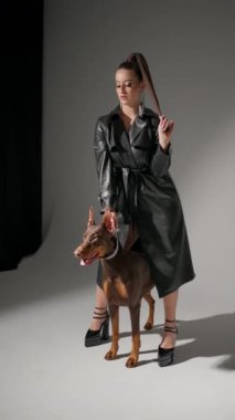 Siyah deri pelerinli kadın Doberman 'ın yanında duruyor. Bir tutam saçı var ve köpek parlak bir tasma takıyor. Arka plan gridir, sahnenin stilini ve zarafetini vurgular..