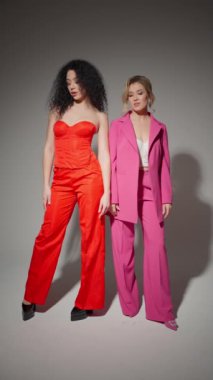 İki genç bayan model parlak takımlar içinde poz veriyor. Biri kırmızı korse ve pantolon giymiş, diğeri de pembe ceket giymiş..