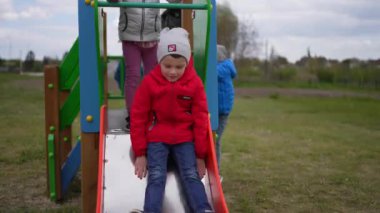 Kırmızı ceketli ve gri şapkalı çocuk bulutlu bir günde çocuk parkında kayıyor. Diğer çocuklar sıralarını bekliyorlar..