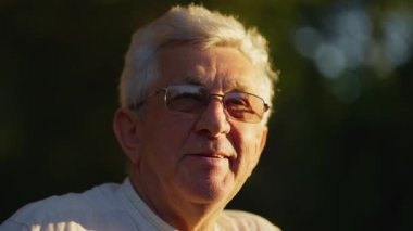 Gri saçlı ve gözlüklü yaşlı adam doğada duruyor. Yüzü, hoş ve sakin bir atmosfer yaratan sıcak güneş ışığıyla aydınlanır..