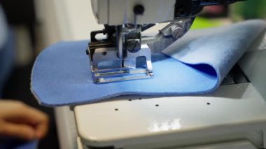 Dikiş makinesi mavi kumaş dikiyor. Modern dikiş ekipmanları hassas ve yüksek kaliteli dikişler sağlar. Dikiş işlemi profesyonel ortamda gerçekleşir.