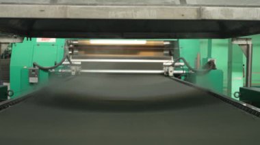 Tekstil fabrikasındaki makine endüstriyel süreçte kumaşı işliyor. Geniş taşıma bandı kumaşı üretimin çeşitli aşamalarında hareket ettirir.