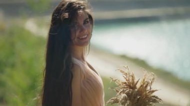 Arkaplan olarak parlayan gölle, açık bej elbiseli kadın ışıldıyor, çekici bir gülümseme, saçları hafifçe yüzünü çerçeveliyor..