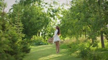 Beyaz elbiseli ve şapkalı kadın ağaçların ve çalıların arasında yeşil çimlerde yalınayak yürüyor. Parlak yaz güneşi yemyeşil bahçeyi aydınlatıyor..