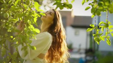 Uzun saçlı bir kadın ağaçların arasında duruyor, sıcak güneş ışınlarıyla yeşillik kokusu soluyor..