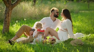 Çocuklu aile güneşli bir günde çimenlerde pikniğin tadını çıkarıyor. Çift yeşil çimenlerde otururken öpüşüyor, çocuk bir parça karpuz tutuyor, yiyecek sepeti yakınlarda..