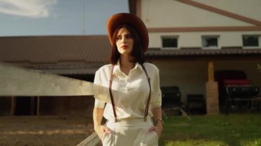 Beyaz elbiseli ve kahverengi şapkalı genç bir kadın arka planda çiftlik binası olan çitlerin yanında duruyor. Ellerini cebinde tutar ve uzaklara bakar..