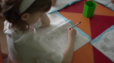 Anaokulu sınıfında, kız çizime odaklanıyor, elindeki kalem kağıda basit egzersizlerle dokunuyor..