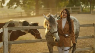 Gün batımında çiftlikte atın yanında duran zarif bir kadın. Kahverengi ceket ve beyaz pantolon giyiyor..