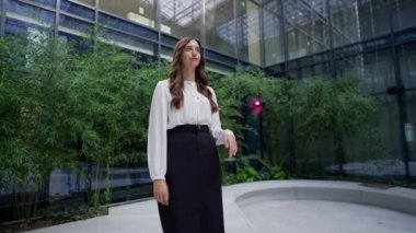 Profesyonel giysiler içindeki iş kadını modern mimarinin çerçevelediği ofis avlusunun verimli manzarasında güven içinde duruyor..