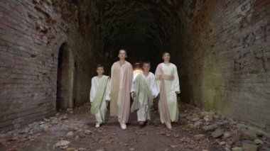 Dönem kostümleri giymiş bir grup çocuk tuğla duvarlar ve kemerlerle antik zindanlarda yürüyorlar. Çocuk kıyafetleri eski çağlara göre stilize edilir..