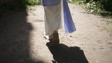 Omuzlarında mavi kefen olan açık renk bornozlu genç bir kadın bina harabelerinin yanındaki yolda ilerliyor. Güneş ışığı onu arkadan aydınlatıyor.
