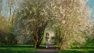 Güneş gözlüklü kadın, bahar parkında çiçek açan ağaçların arasında asfalt yolda koşuyor. Beyaz çiçekli ağaçlar yol üzerinde kemer oluşturur..
